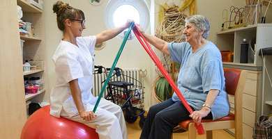 Foto: Eine Therapeutin trainiert mit Hilfe von Bändern mit einer Patietin ihre Beweglichkeit